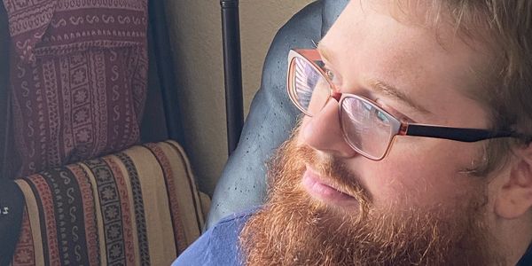 red bearded man sitting in black recliner in front of lamp and baby wraps. he is wearing glasses