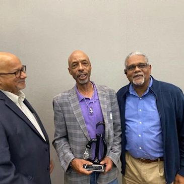 Three smiling men standing together, one holding an award.