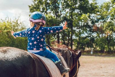 A child riding a horse with their arms extended out. The child is wearing a helmet and a star shirt. 