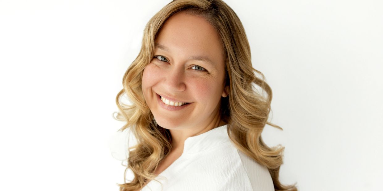 Smiling woman with wavy blonde hair wearing a white top against a white background.