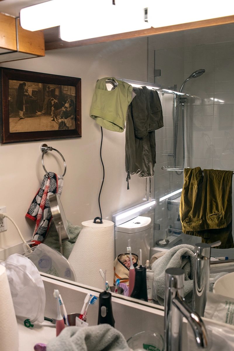 A cluttered bathroom with clothes hanging and toiletries on the sink.