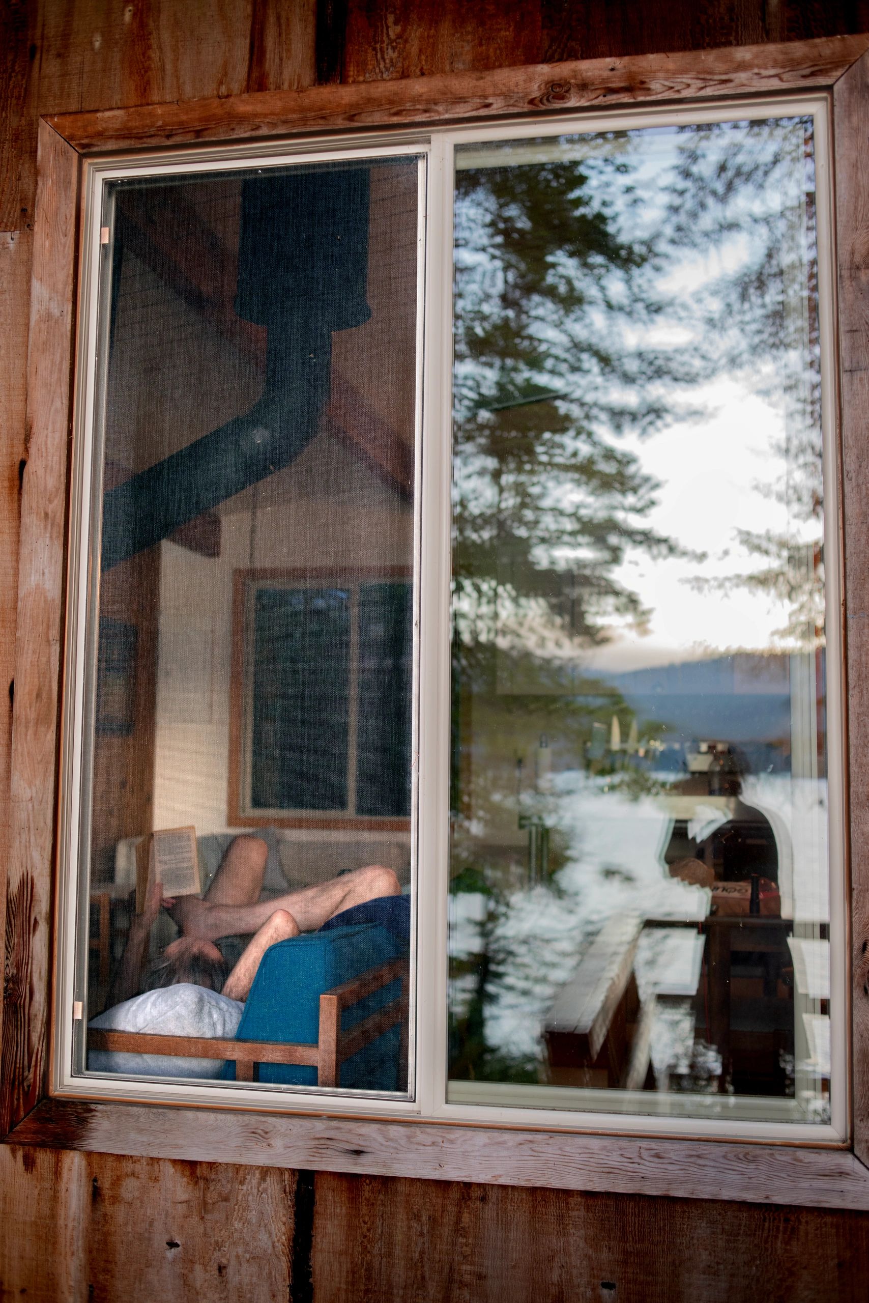 Person reading while lounging on a chair, seen through a window with reflections.