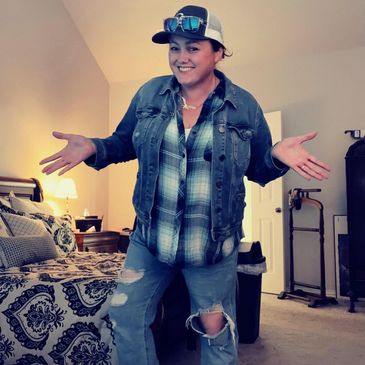 Person posing casually in a cozy bedroom wearing a denim jacket and ripped jeans.