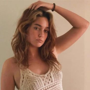 A woman with wavy hair poses with her hand on her head wearing a crochet top.