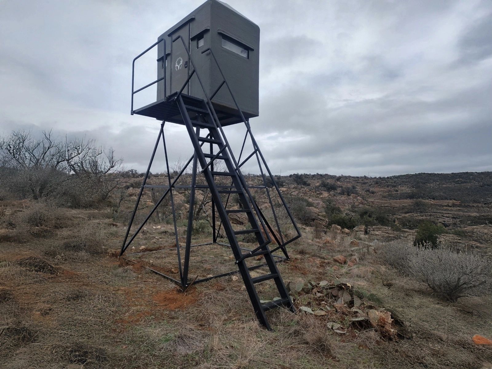 A black hunting blind elevated on metal legs in a dry, rocky landscape under a cloudy sky.
