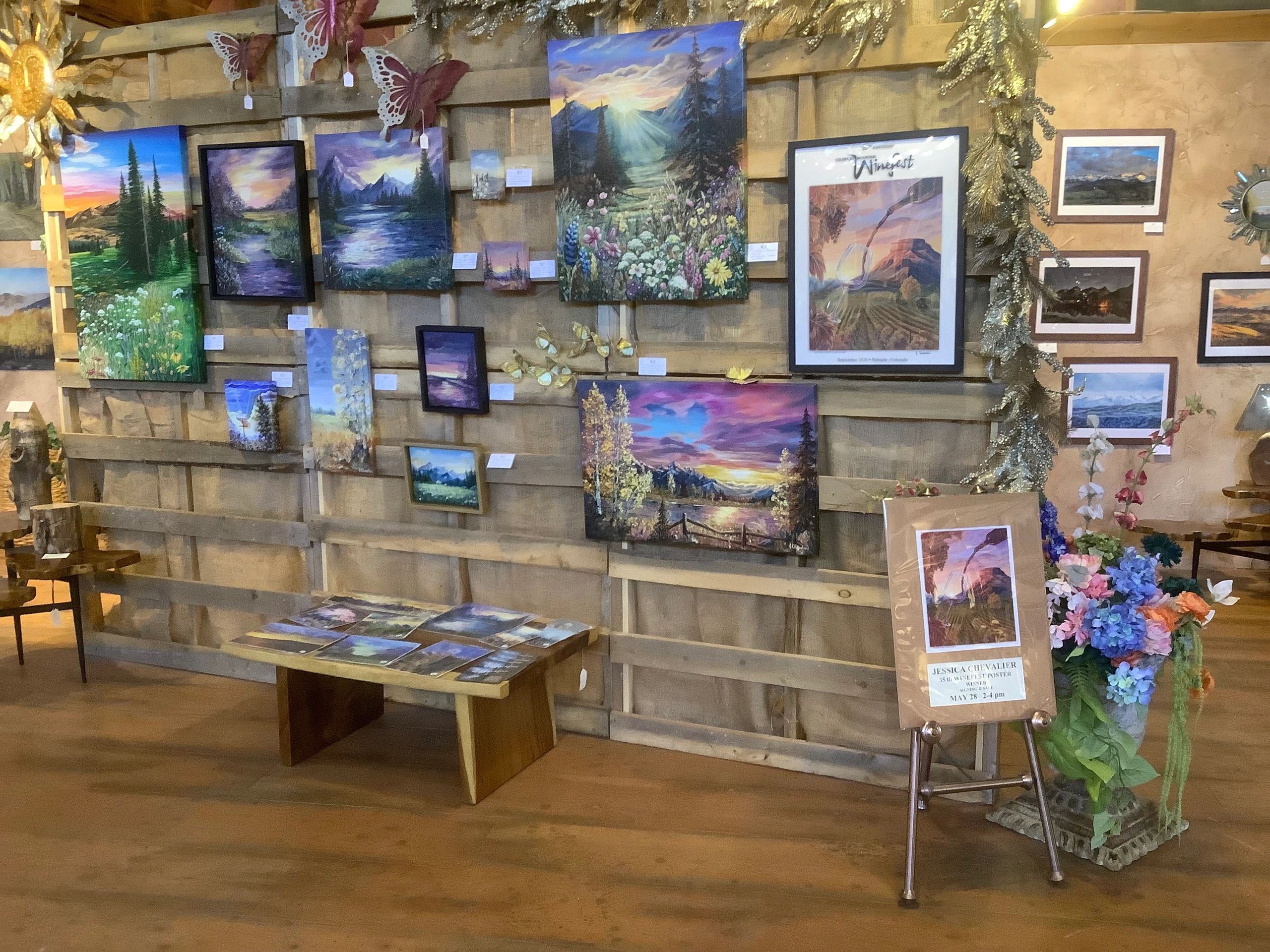 Gallery wall displaying colorful landscape and nature paintings with flowers and butterflies.