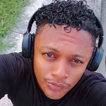 Man with curly hair wearing headphones and a black shirt taking a selfie outdoors.