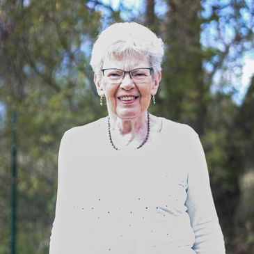 Smiling elderly woman with glasses outdoors in a white top.