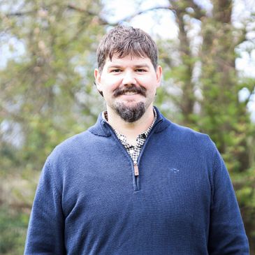 Smiling man in a blue sweater standing outdoors with trees in the background.