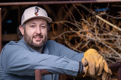 Man in work gloves and cap leaning on metal railing with dried branches in background.