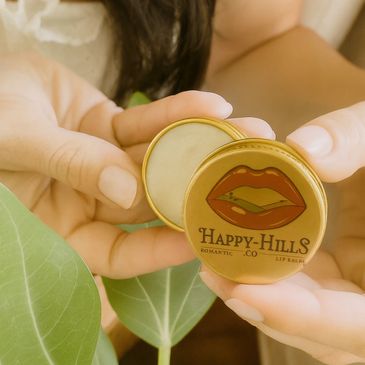A person holds open a tin of Happy Hills lip balm near green leaves.