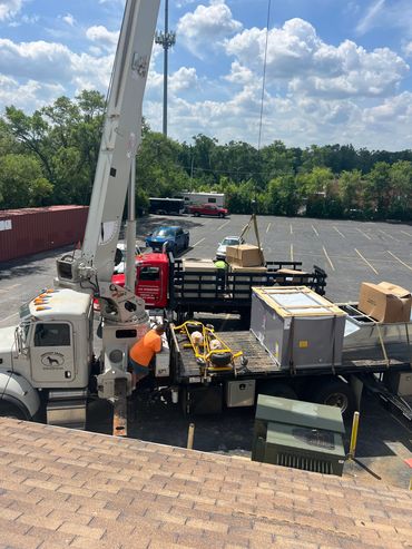Workers loading heavy equipment onto a flatbed truck with a crane in a parking lot.