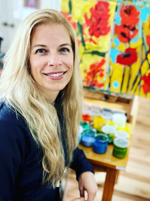 Smiling woman with long blonde hair in front of colorful painting and art supplies.