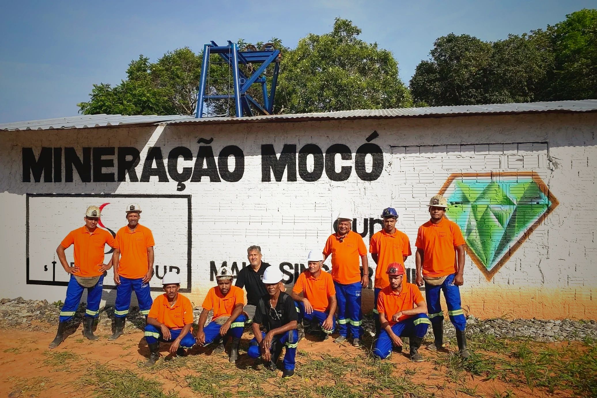 Group of miners in orange shirts posing in front of a mining company wall.