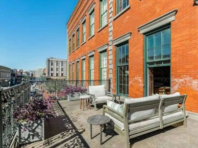 Sunny balcony with cozy seating and city views beside a red brick building.