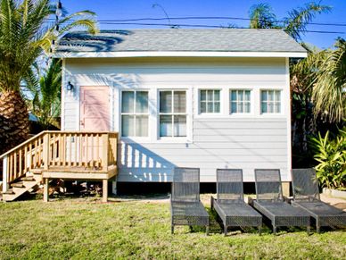 Small white beach house with four black lounge chairs on lawn.