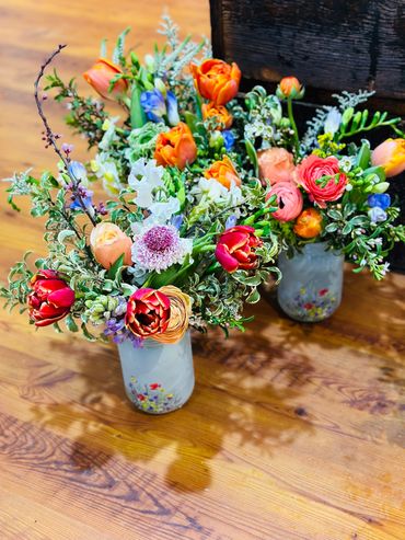 Two vibrant flower bouquets in decorative jars on a wooden floor.