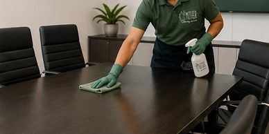 A man cleans a conference room table with gloves and spray.