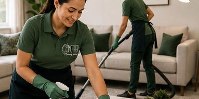Two women in green uniforms cleaning a living room thoroughly.