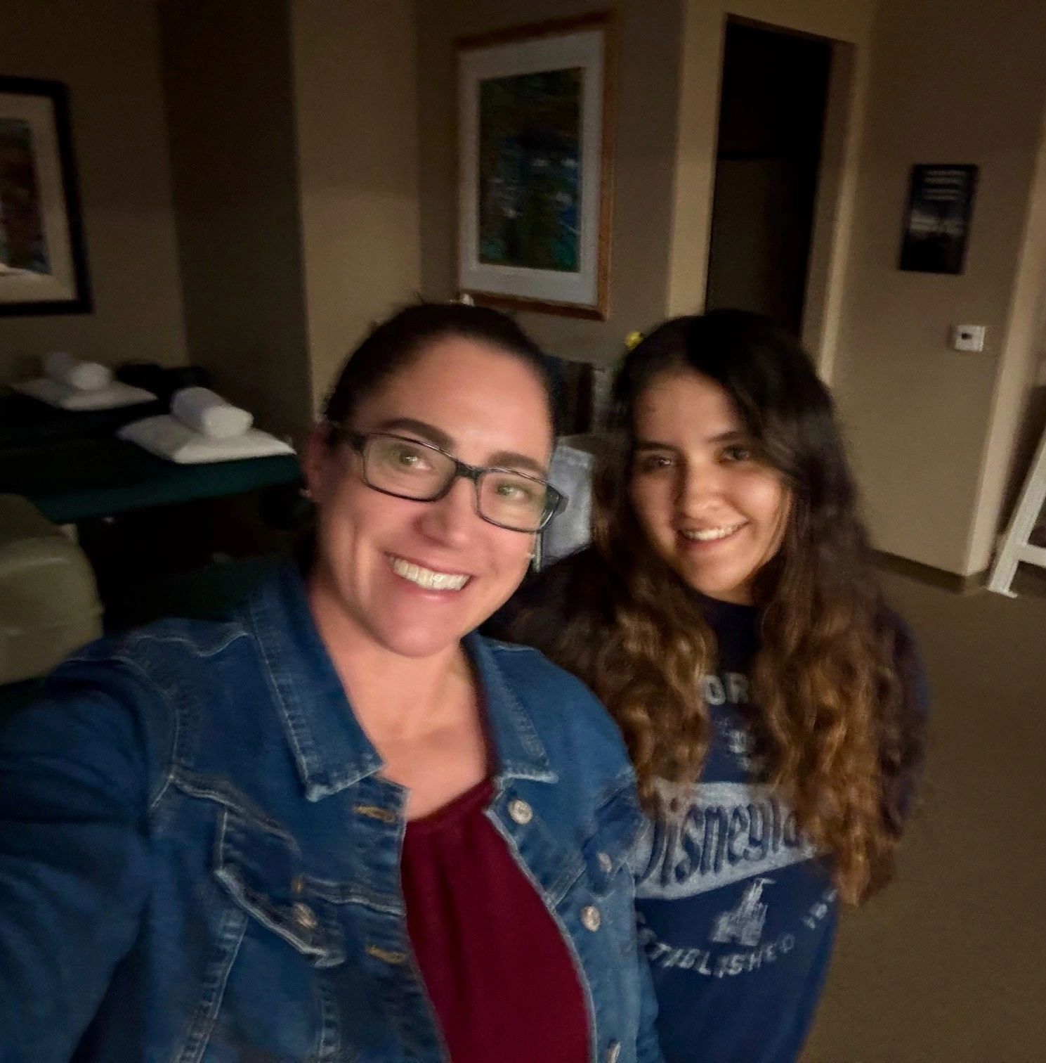 Two women smiling indoors, one wearing glasses and denim jacket.