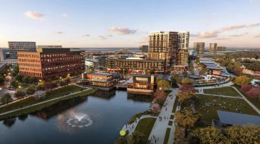 Urban waterfront area with buildings, park, and fountain at sunset.