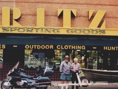 Two men stand outside Ritz Sporting Goods store with motorcycles and a boat.
