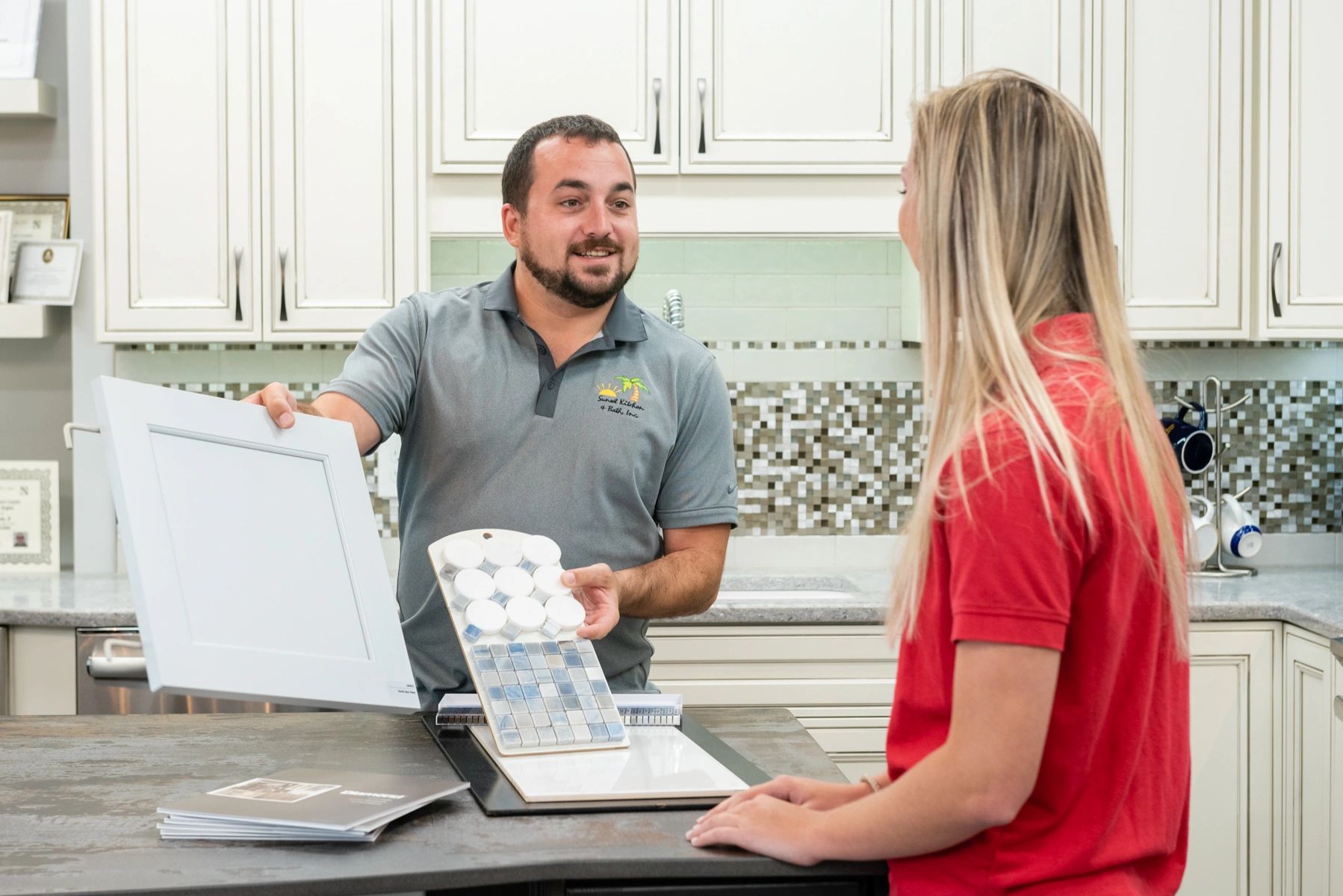 Man shows cabinet door and tile samples to woman in kitchen.