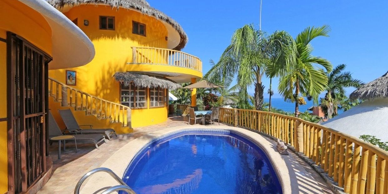 A vibrant yellow house with a thatched roof beside a blue pool and tropical palm trees.