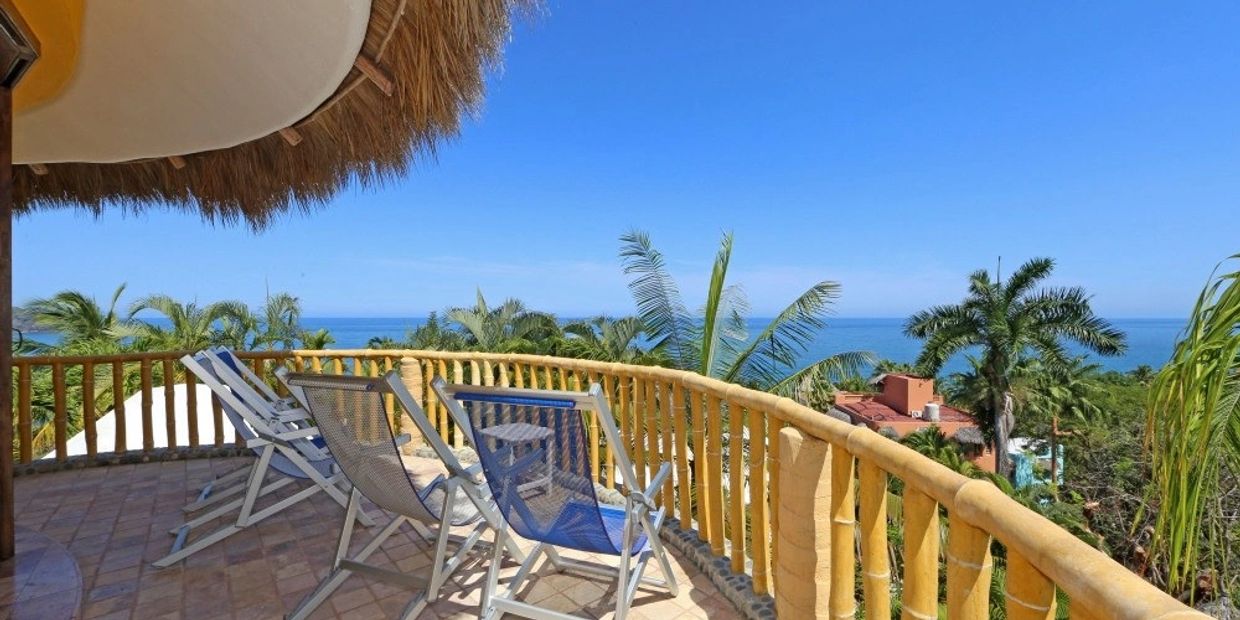 Balcony with chairs overlooking the ocean and palm trees under a clear blue sky.