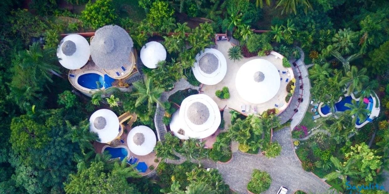 Aerial view of a tropical resort with round thatched-roof huts and pools.