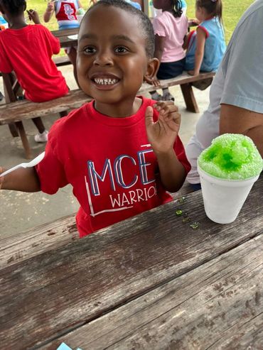 Smiling child in a red shirt enjoying a green shaved ice treat outdoors.