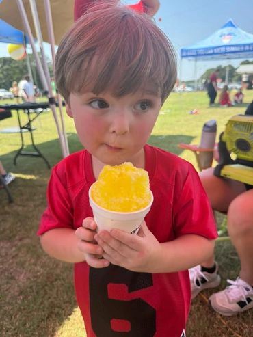 Young boy holding a cup of yellow shaved ice at an outdoor event.