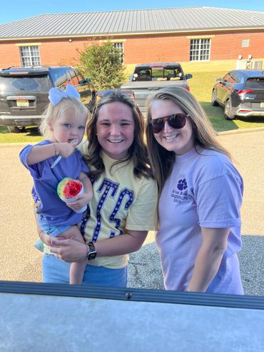 Three smiling individuals, including a child eating a colorful snow cone, standing outdoors.