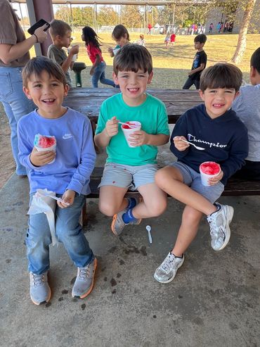 Three boys happily eating shaved ice while sitting on a bench outdoors.