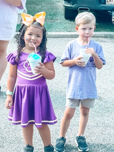 Two children enjoying blue shaved ice, one in a purple dress with a big yellow bow.