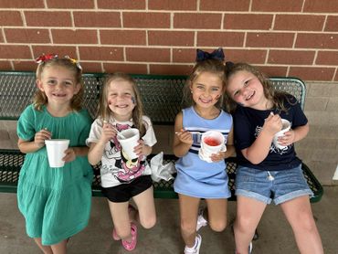 Four young girls sitting on a bench eating snow cones and smiling.