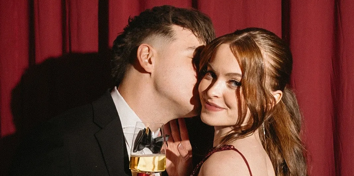 A man whispers to a smiling woman holding a drink, against a red curtain background.