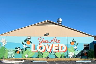 A mural on a building says 'You Are Loved' with floral and fruit designs.