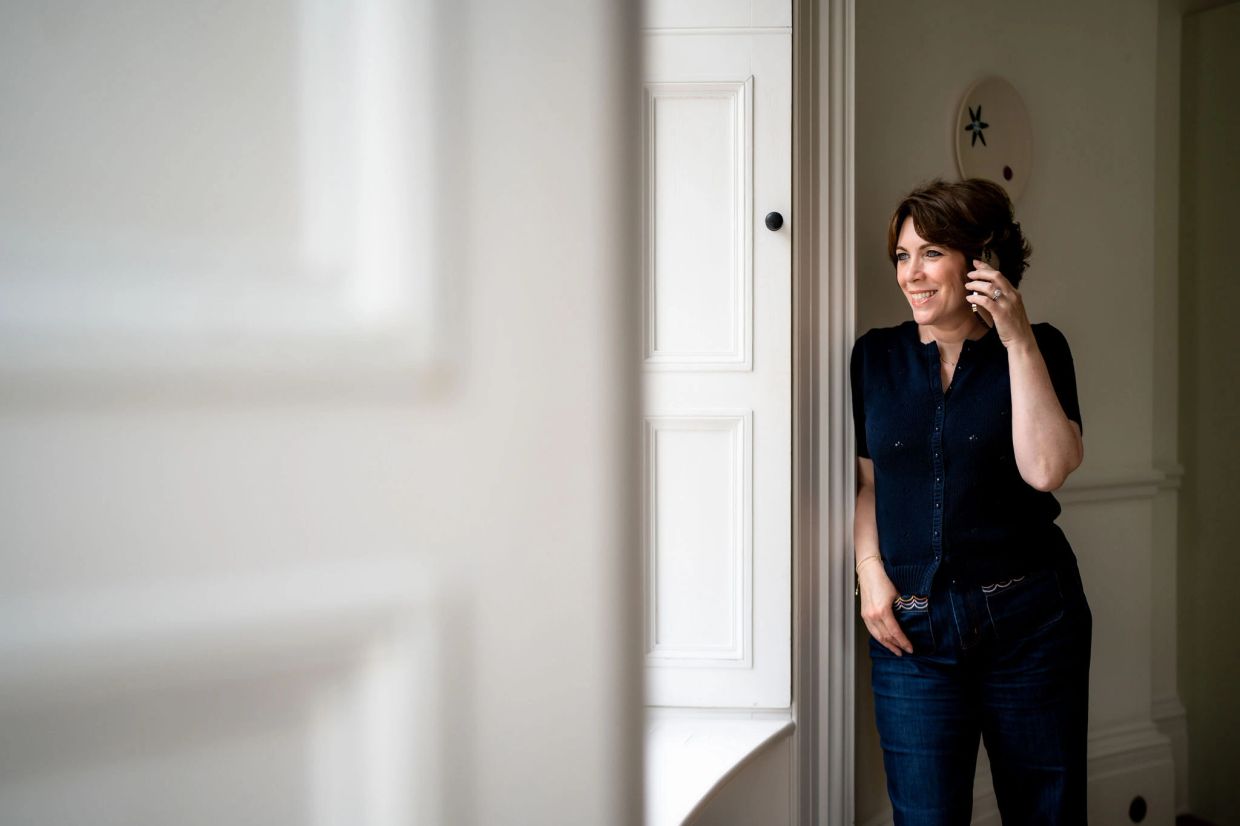 Woman smiling and talking on phone, standing by a window in a cozy room.