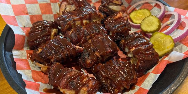BBQ pork rib tips drizzled with BBQ sauce served on a picnic plater with dill pickles and red onion.