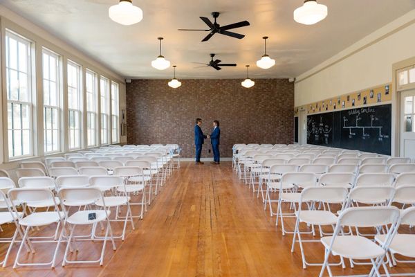 Two men in blue suits shake hands in a bright, empty room with white chairs.