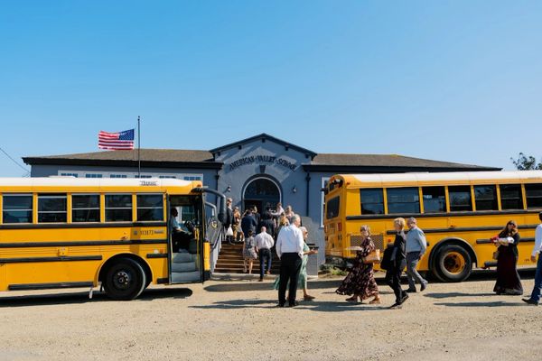 People gather outside American Valley School near two yellow school buses on a sunny day.