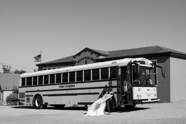 Newlyweds share a romantic kiss beside a school bus in front of a building.