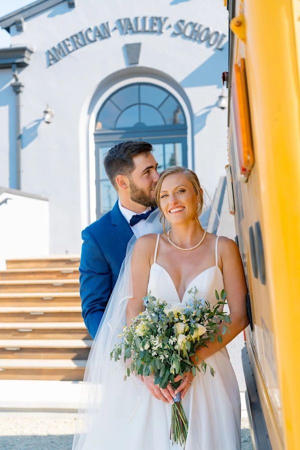 A groom kisses his bride on the cheek outside American Valley School, both dressed in wedding attire.