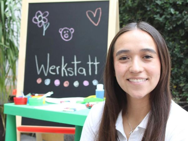 Smiling woman in front of a chalkboard with 'Werkstatt' and colorful drawings.