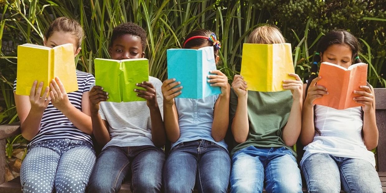 Five children sitting on a bench, each reading a colorful book outdoors.