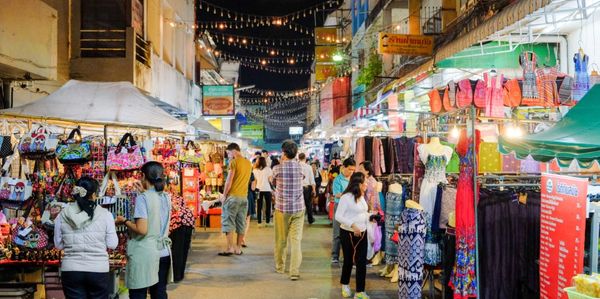 Night market street with colorful stalls and shoppers under string lights.
