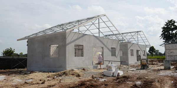 Partially constructed houses with metal roof frames on a dirt site under a cloudy sky.