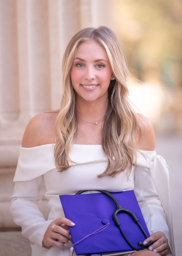 Young woman in white dress holding graduation cap with stethoscope.