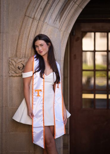 Young woman in white dress and graduation stole leaning on stone arch.